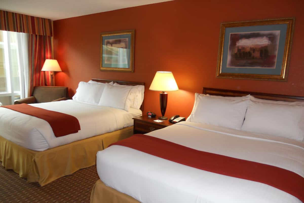 A hotel room with two beds, each with white linens and red throw blankets, a wooden headboard, a nightstand with a lamp, and a framed picture on the wall.