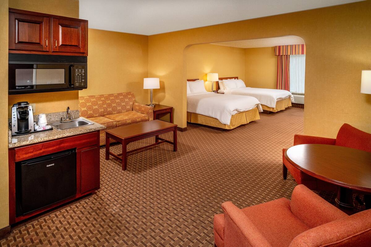 A hotel room with a kitchenette, two beds, a sofa, a dining table, and a carpeted floor.