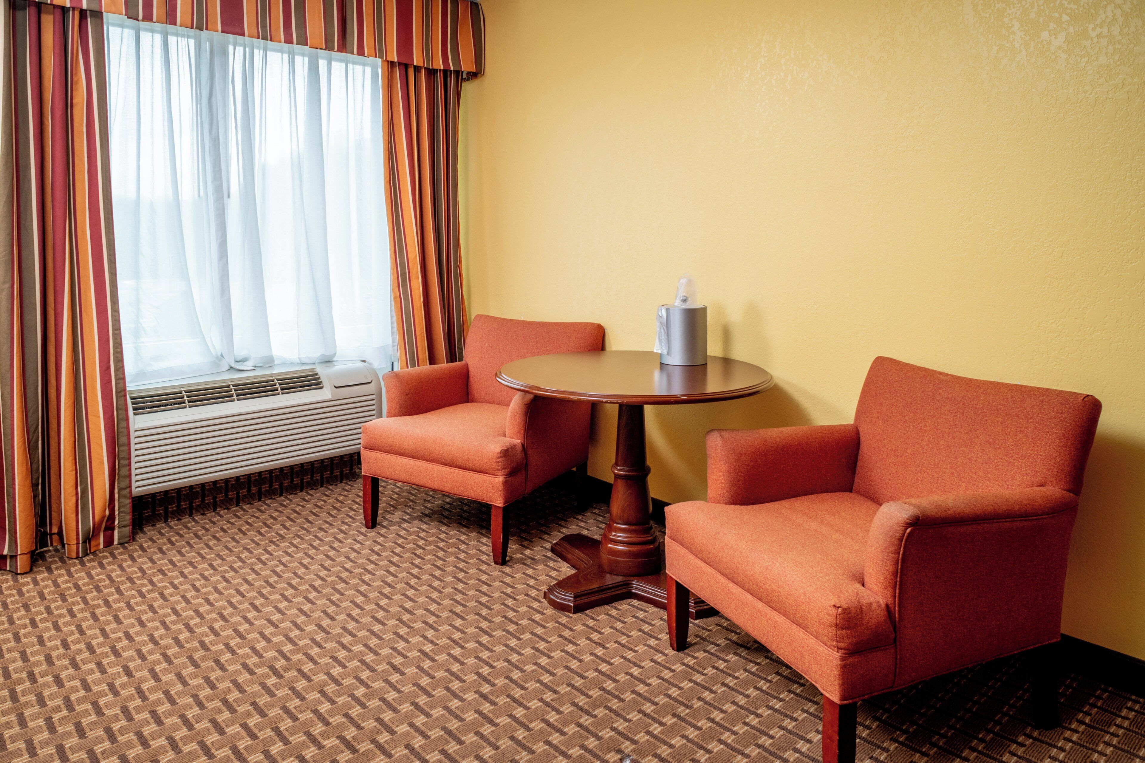 Quarto de hotel com duas poltronas, uma mesinha redonda, janela com cortinas e aparelho de ar condicionado.