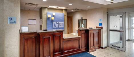 A hotel reception area with a wooden reception desk, a blue carpet, and a glass elevator.
