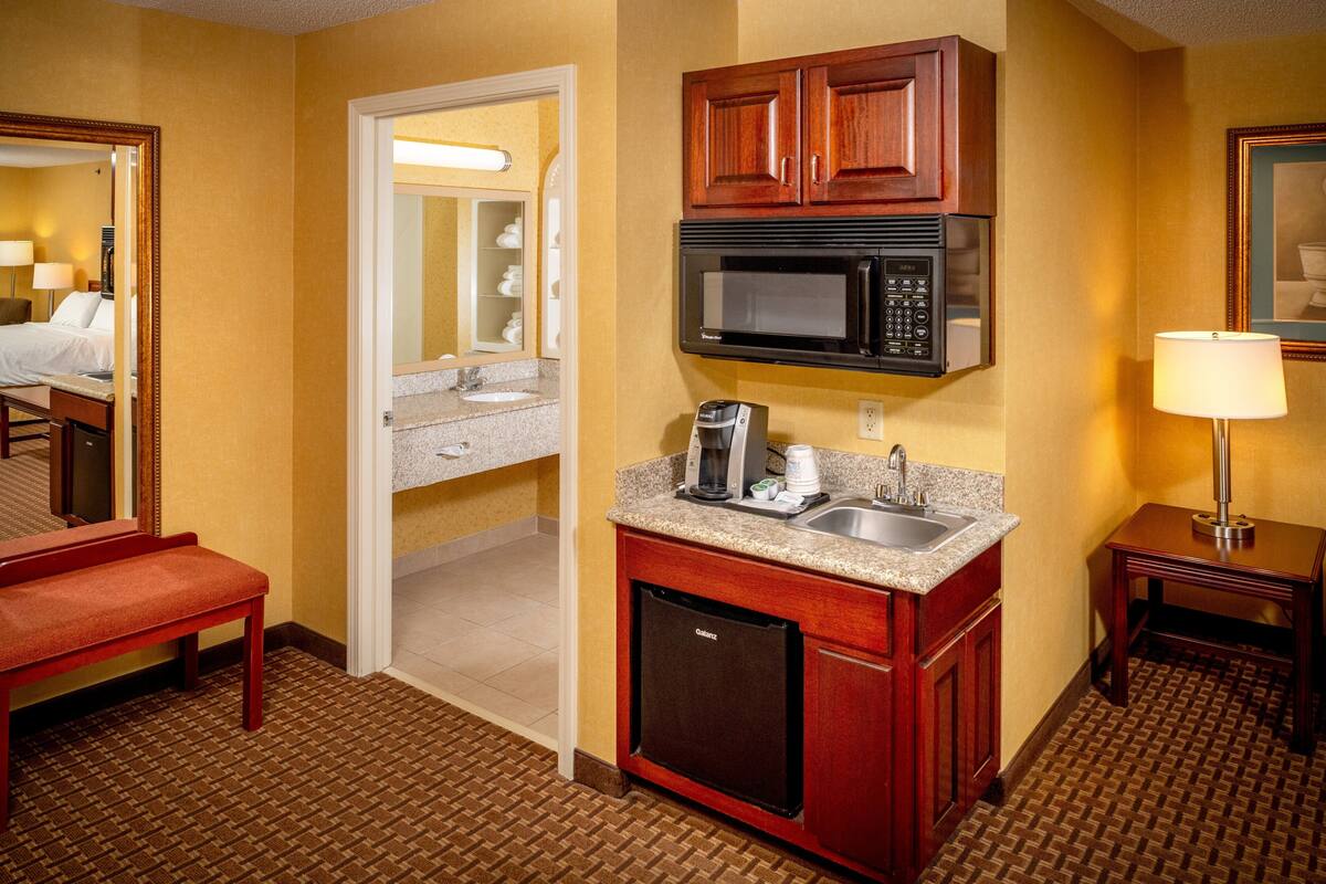 A hotel room with a kitchenette, a microwave, a coffee maker, a sink, and a bench.