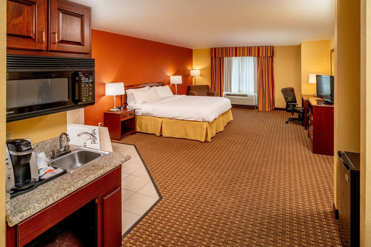 A hotel room with a bed, a desk, a TV, a microwave, a sink, and a window with curtains.