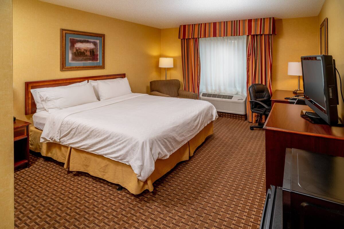 A hotel room with a large bed, a desk with a television, a chair, and an air conditioning unit.
