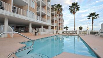 A swimming pool with a metal handrail, surrounded by a building with balconies and palm trees.