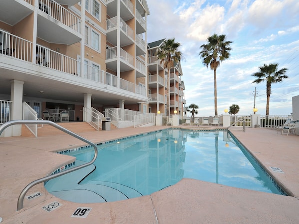 A swimming pool with a metal handrail, surrounded by a building with balconies and palm trees.