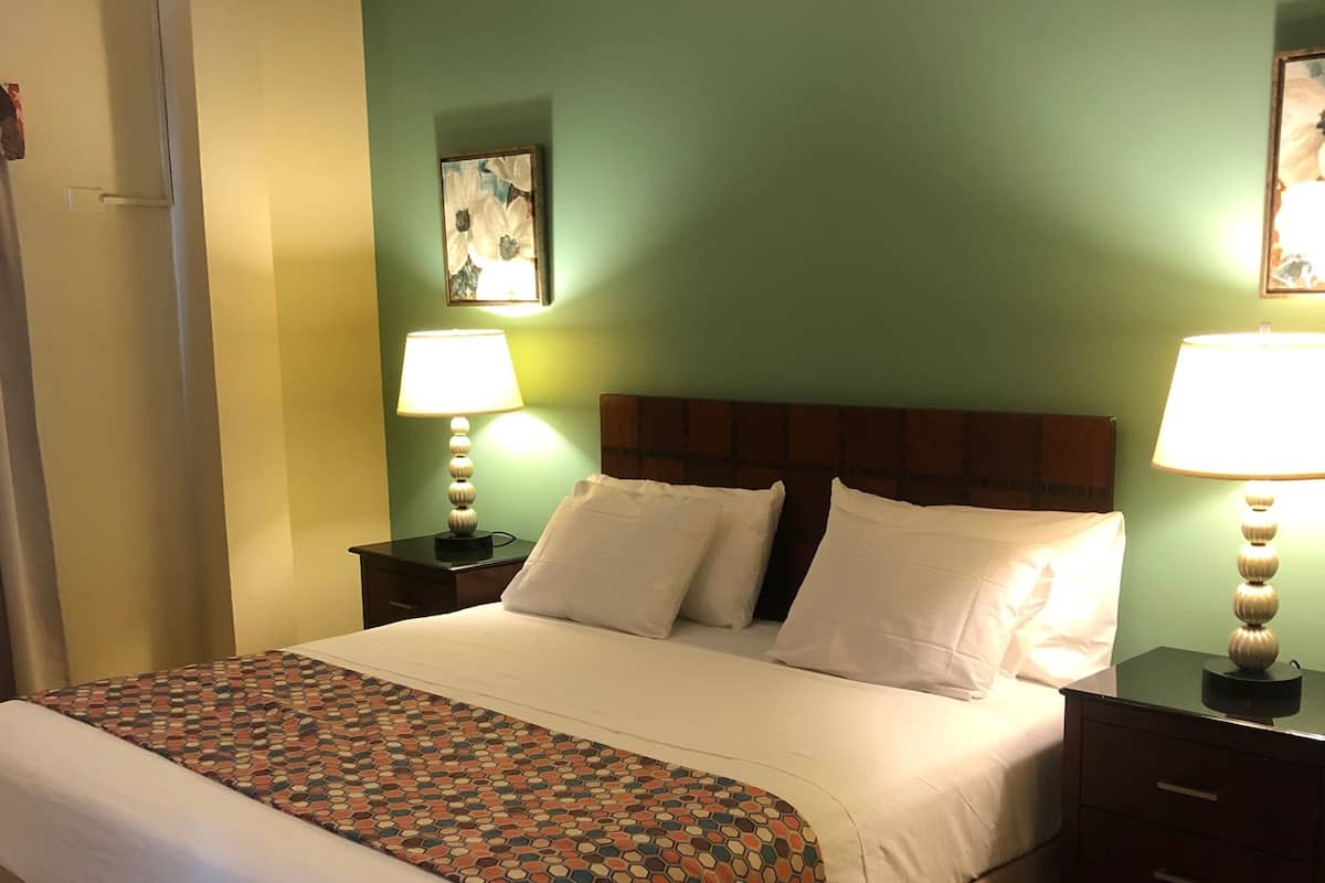 A hotel room with a large bed, two bedside tables with lamps, a patterned throw blanket, and a green wall with a painting.