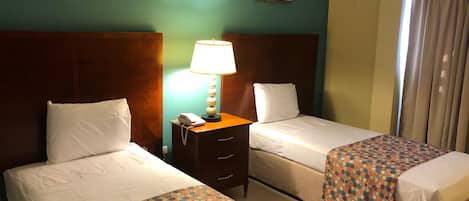 Quarto de hotel com duas camas, uma mesa de cabeceira com abajur e uma pequena mesa.