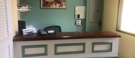A reception desk with a 'NO SMOKING' sign, a phone, and a card holder. There is a painting on the wall and a fire extinguisher on a shelf.