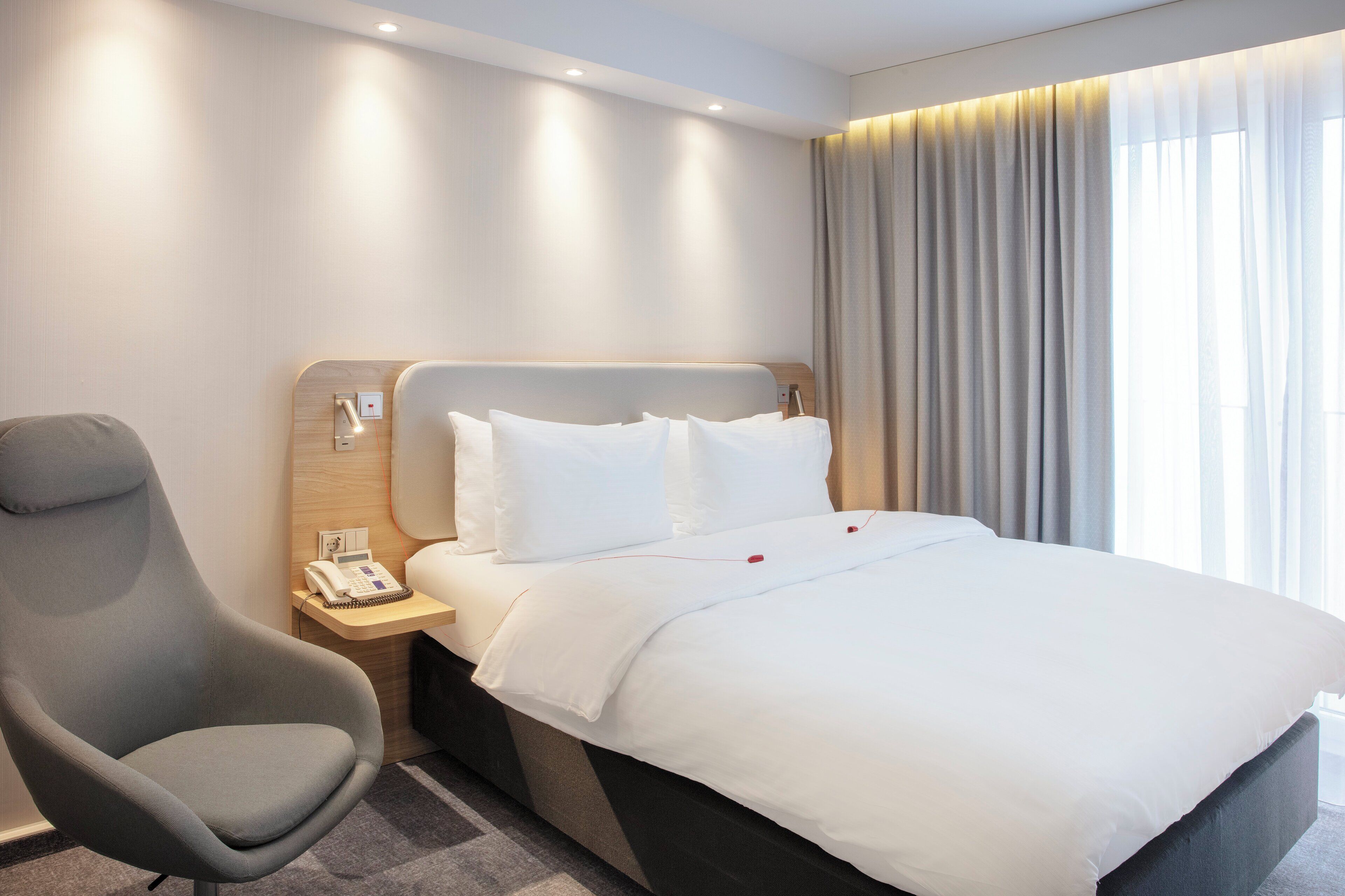 A hotel room with a bed, a chair, a bedside table with a phone, and a window with curtains.