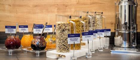 Variedad de alimentos y bebidas a la vista, incluyendo frascos de mermelada, jalea de naranja y diferentes tipos de cereales, con señalización clara que indica sus nombres.