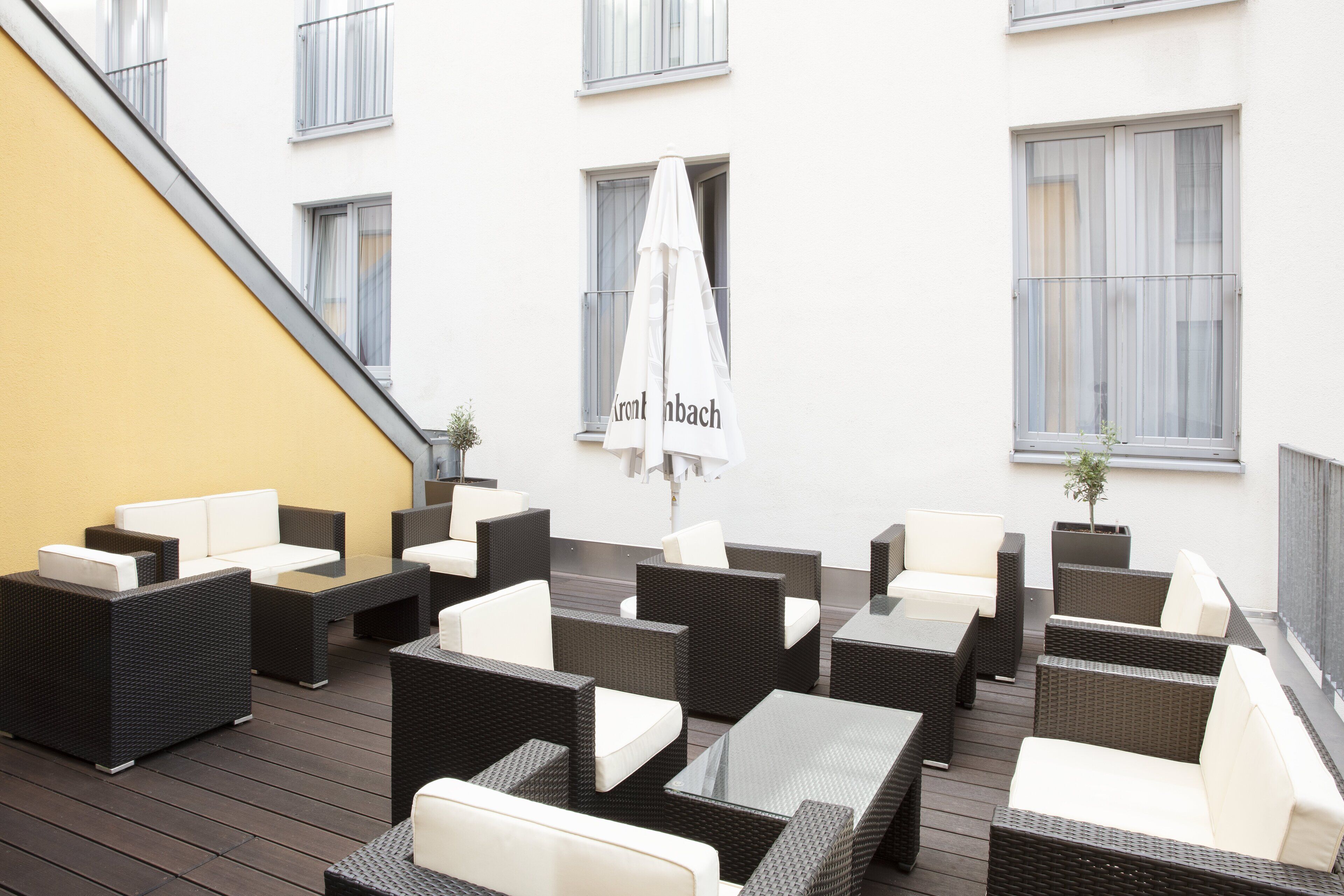 Een terras met rieten meubels en witte kussens, een witte parasol en een gele muur.