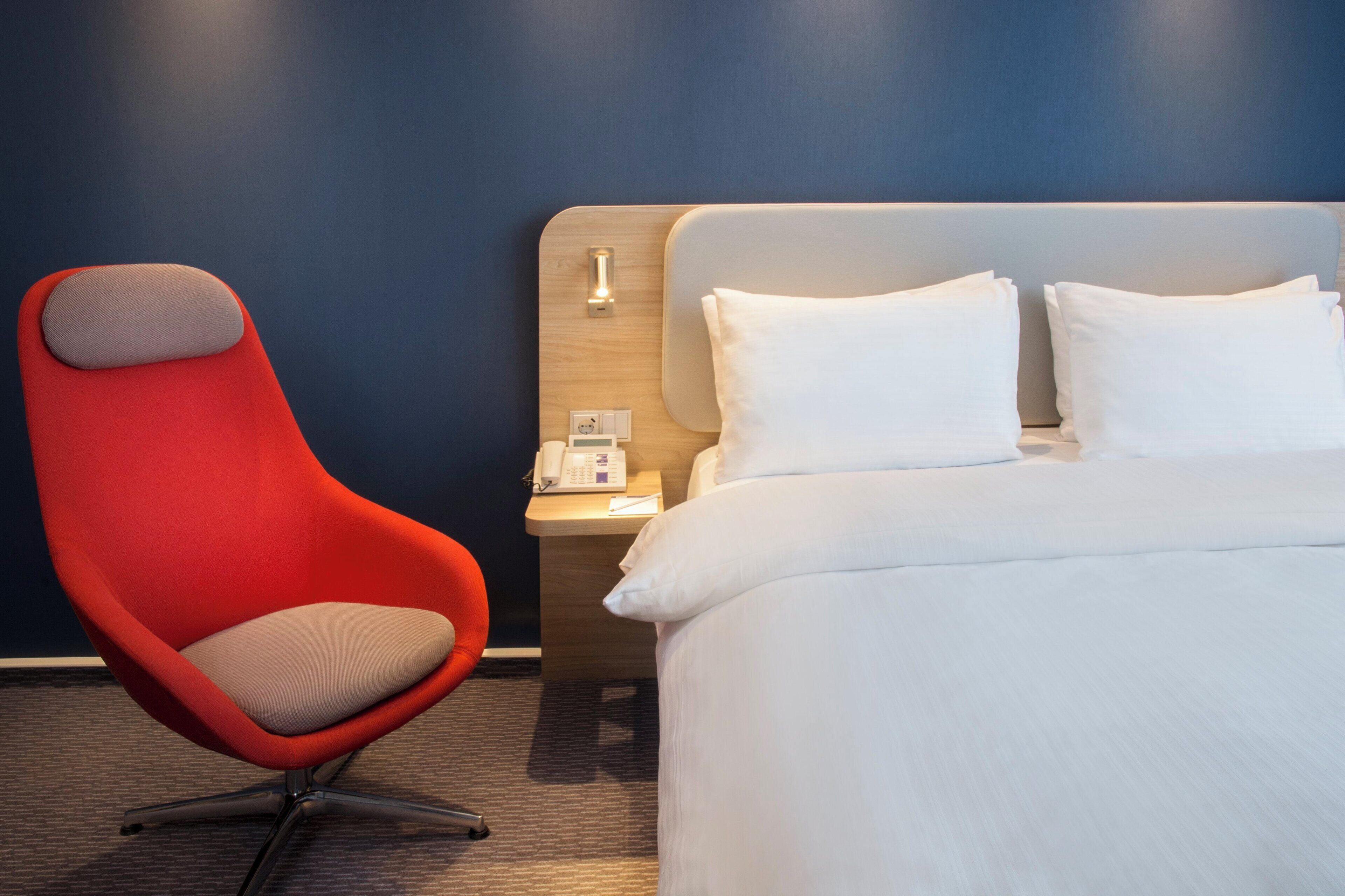 A hotel room with a single bed, a red chair, a bedside table with a lamp, and a wall-mounted phone.