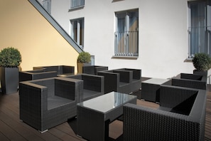 Un espace extérieur aménagé avec des meubles en osier et des tables en verre, entouré de murs et de fenêtres blancs.