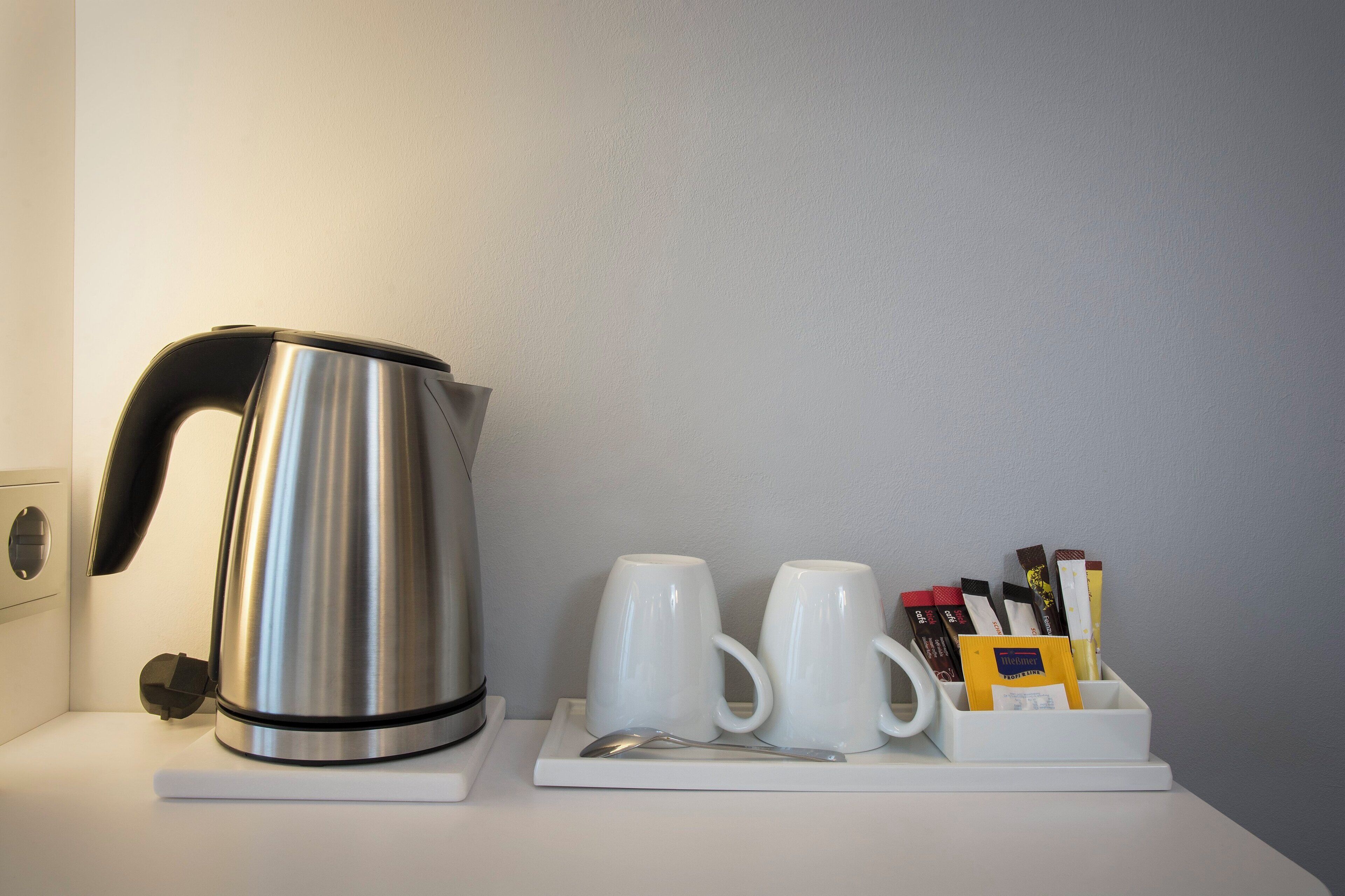 A stainless steel kettle, two white mugs, a spoon, and a tray with coffee packets on a white countertop.