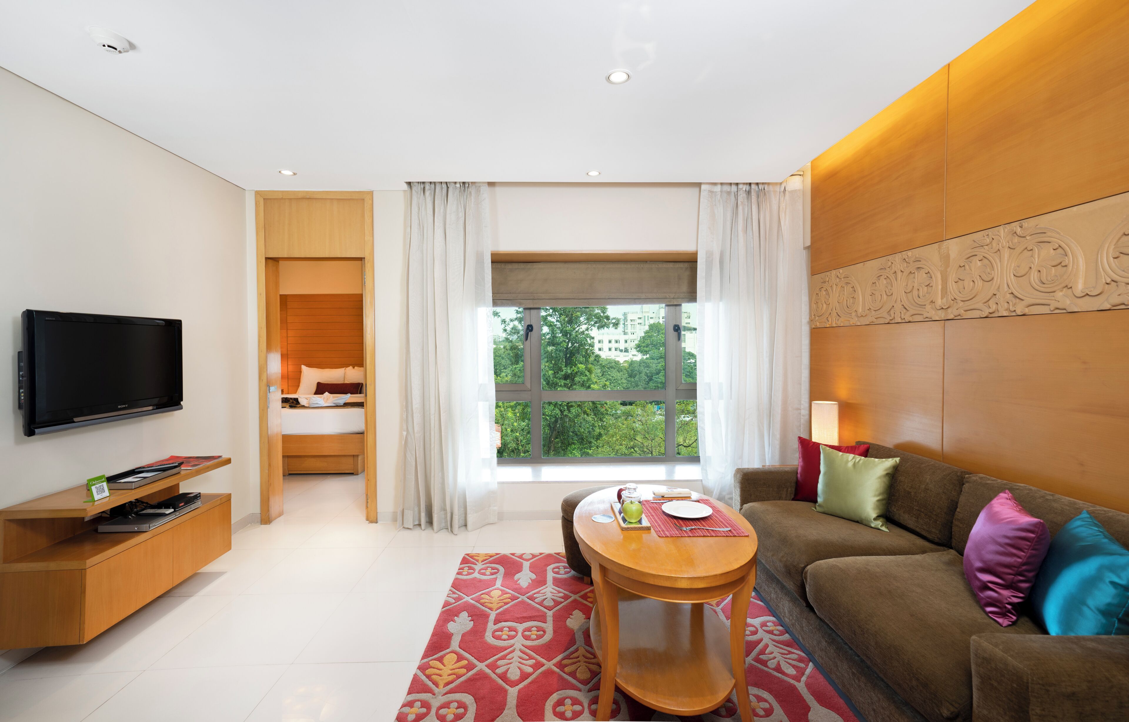 Habitación de hotel moderna con televisión, un mueble bajo de madera, sofá con cojines, una mesita y una ventana con cortinas, y vista a zonas verdes.