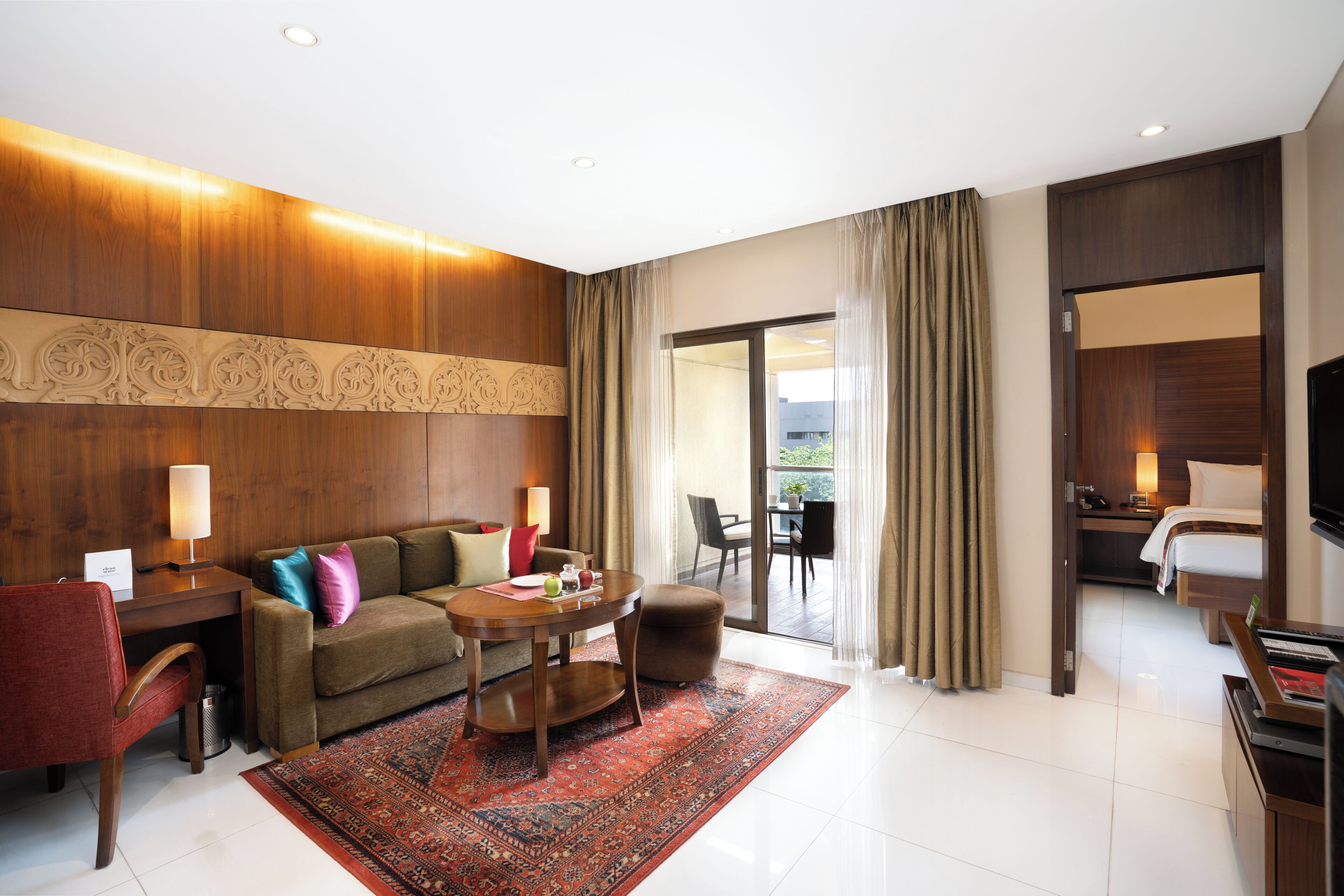 Habitación de hotel con pared de madera decorativa, un sofá, una mesa de centro, un escritorio con lámpara, televisión y balcón con cortinas.