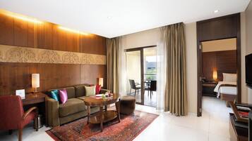 Habitación de hotel con pared de madera decorativa, un sofá, una mesa de centro, un escritorio con lámpara, televisión y balcón con cortinas.