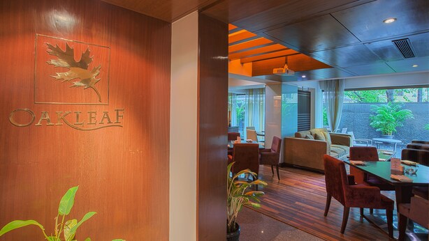 Un restaurante con una pared de madera que presenta un logotipo de hoja y el nombre "Oakleaf". En su interior, hay mesas y sillas de madera, un sofá y un amplio ventanal con vistas al exterior.