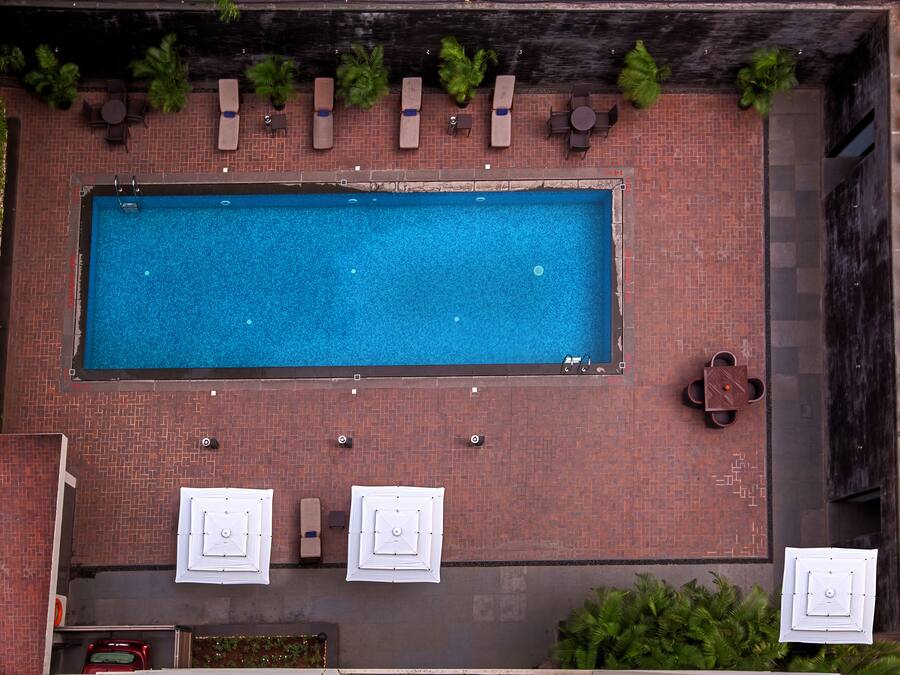 An aerial view of a rectangular swimming pool with lounge chairs and umbrellas, surrounded by a brick patio and greenery.