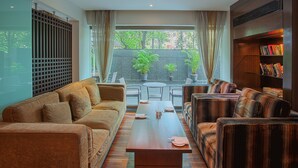 Una sala de estar moderna con un sofá, sillones, una mesa de centro y una estantería. Hay un ventanal con cortinas y vistas al exterior con mucha vegetación.