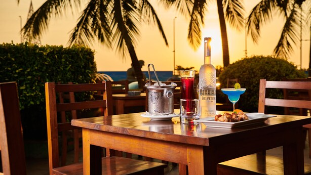 A wooden table with a bottle, a drink, and a plate of food, set against a backdrop of palm trees and a sunset. - Galadari Hotel (Colombo)