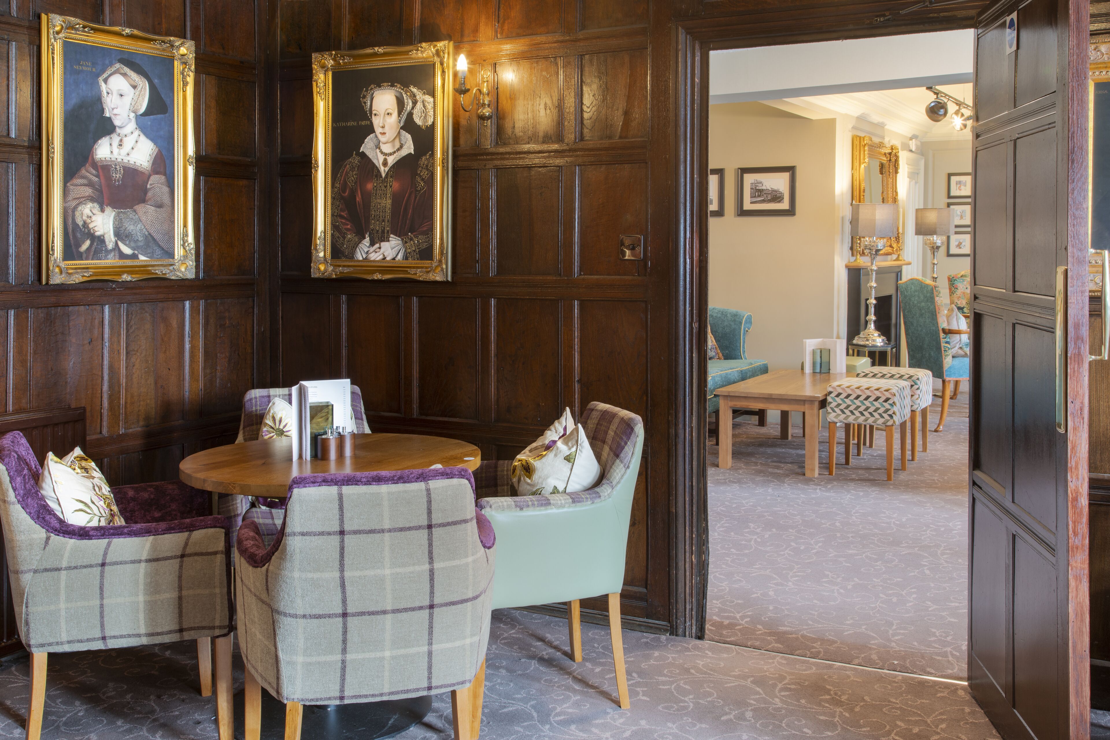 A room with wooden paneled walls, a round wooden table, and two plaid chairs. There are framed portraits on the wall and a door leading to another room.