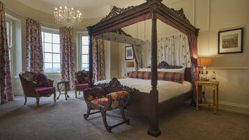 A four-poster canopy bed with a chandelier, floral curtains, and antique furniture in a well-lit room.