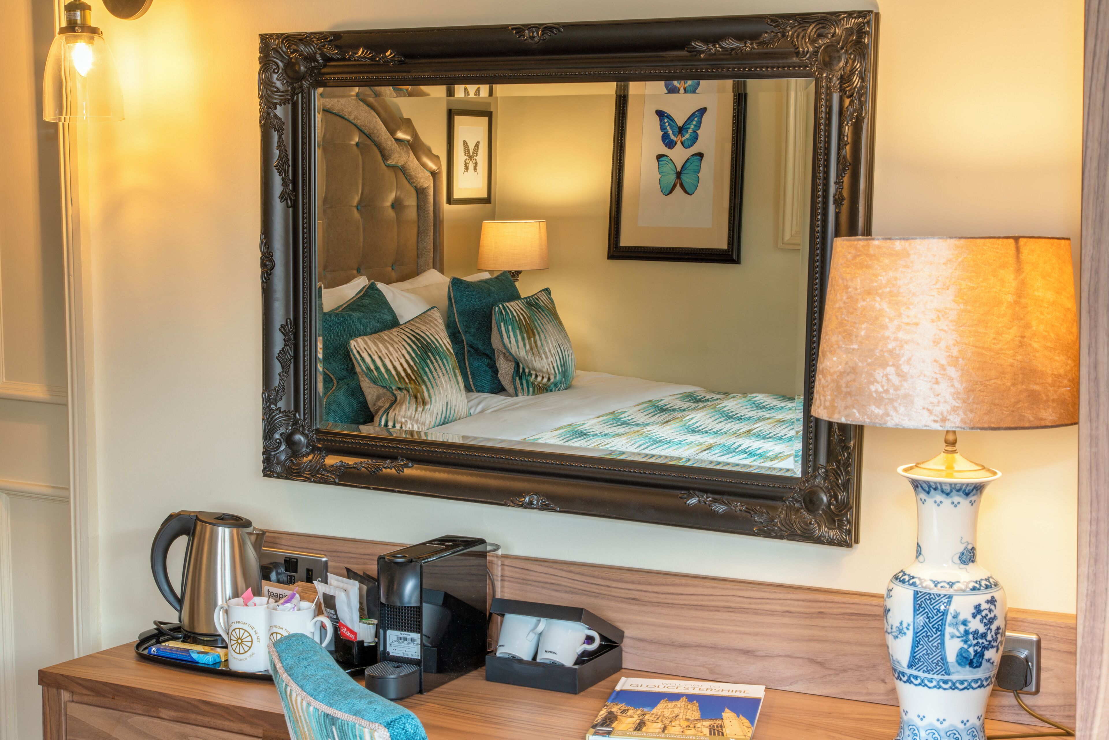 A hotel room with a large mirror, a bedside table with a lamp, a coffee maker, and a framed picture of a building.