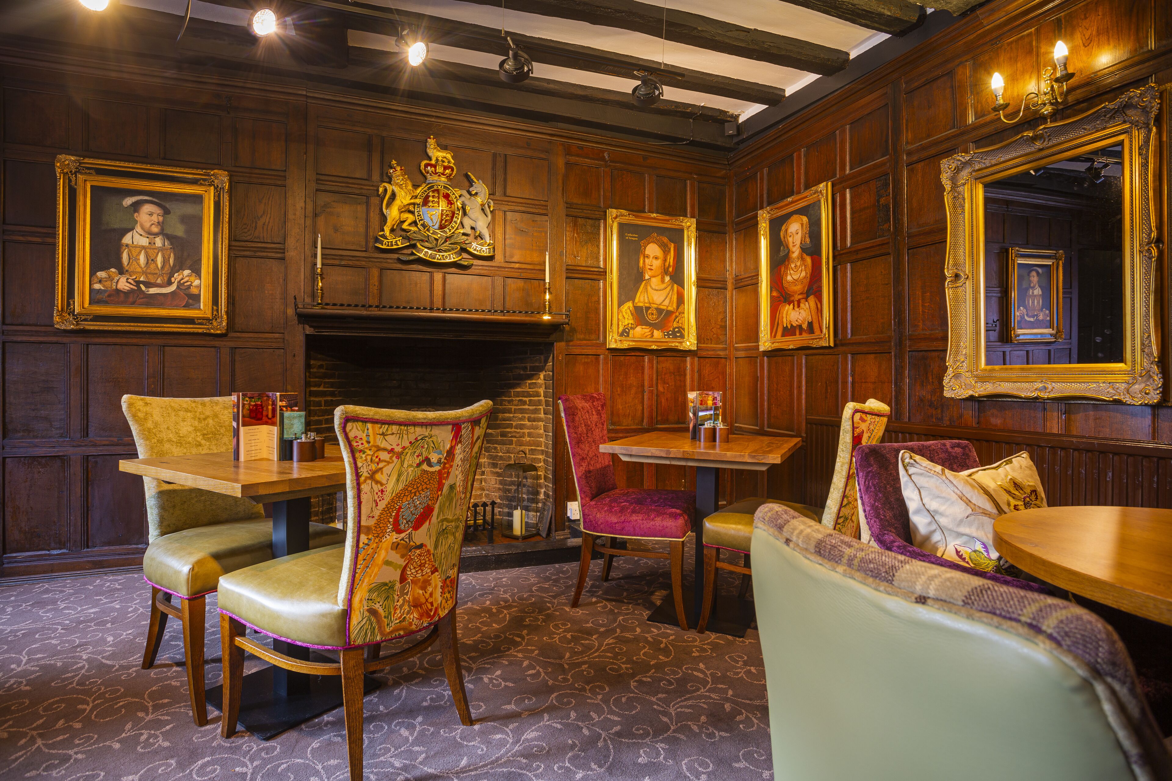 A room with wooden paneled walls, a fireplace, and several framed portraits. There are two dining tables with chairs, one with a purple upholstered chair.