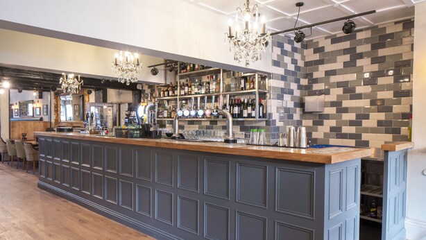 Een bar met een houten toonbank, barstoelen, een kroonluchter en een betegelde achterwand.