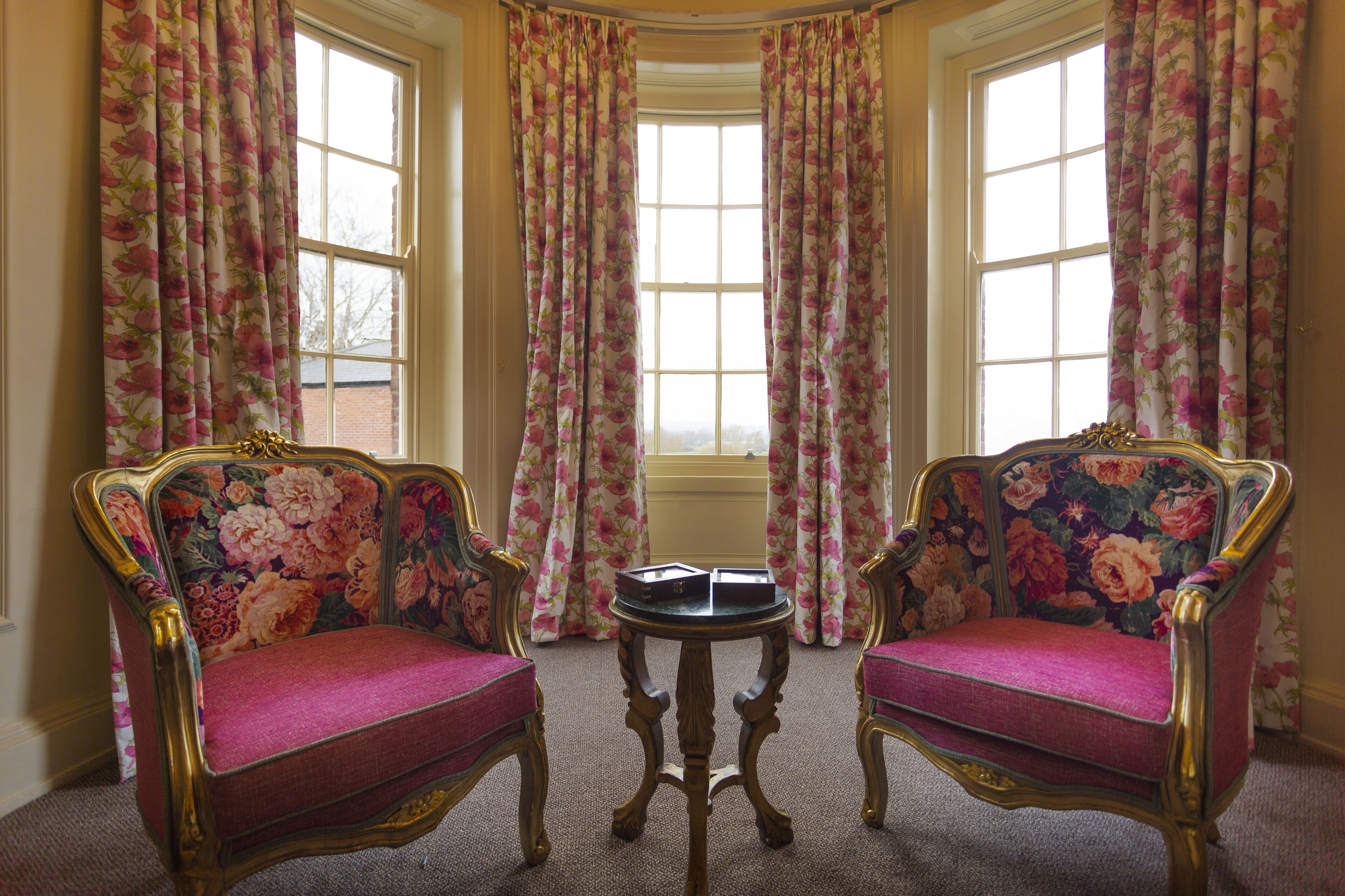 Una habitación con dos sillones ricamente decorados, tapizados con motivos florales, una pequeña mesa redonda y un ventanal con cortinas estampadas.