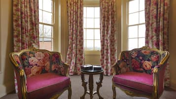Una habitación con dos sillones ricamente decorados, tapizados con motivos florales, una pequeña mesa redonda y un ventanal con cortinas estampadas.