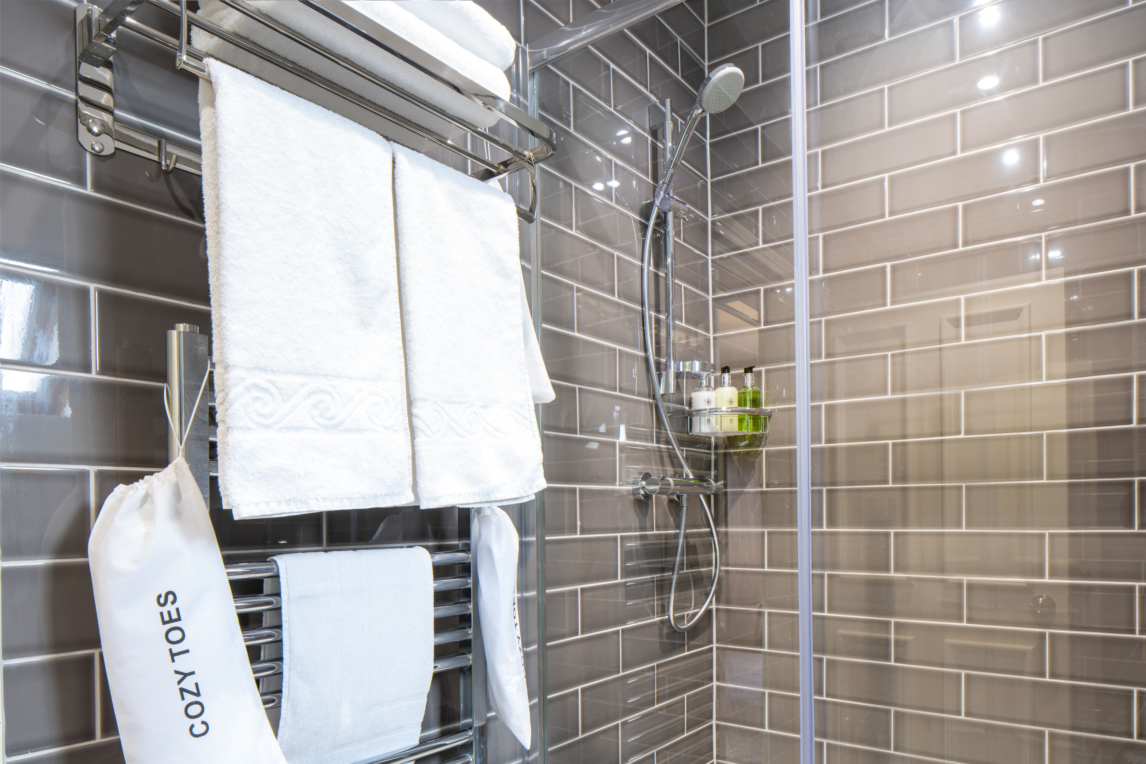 A modern bathroom with grey tiled walls, a glass shower enclosure, and a towel rack holding multiple white towels.