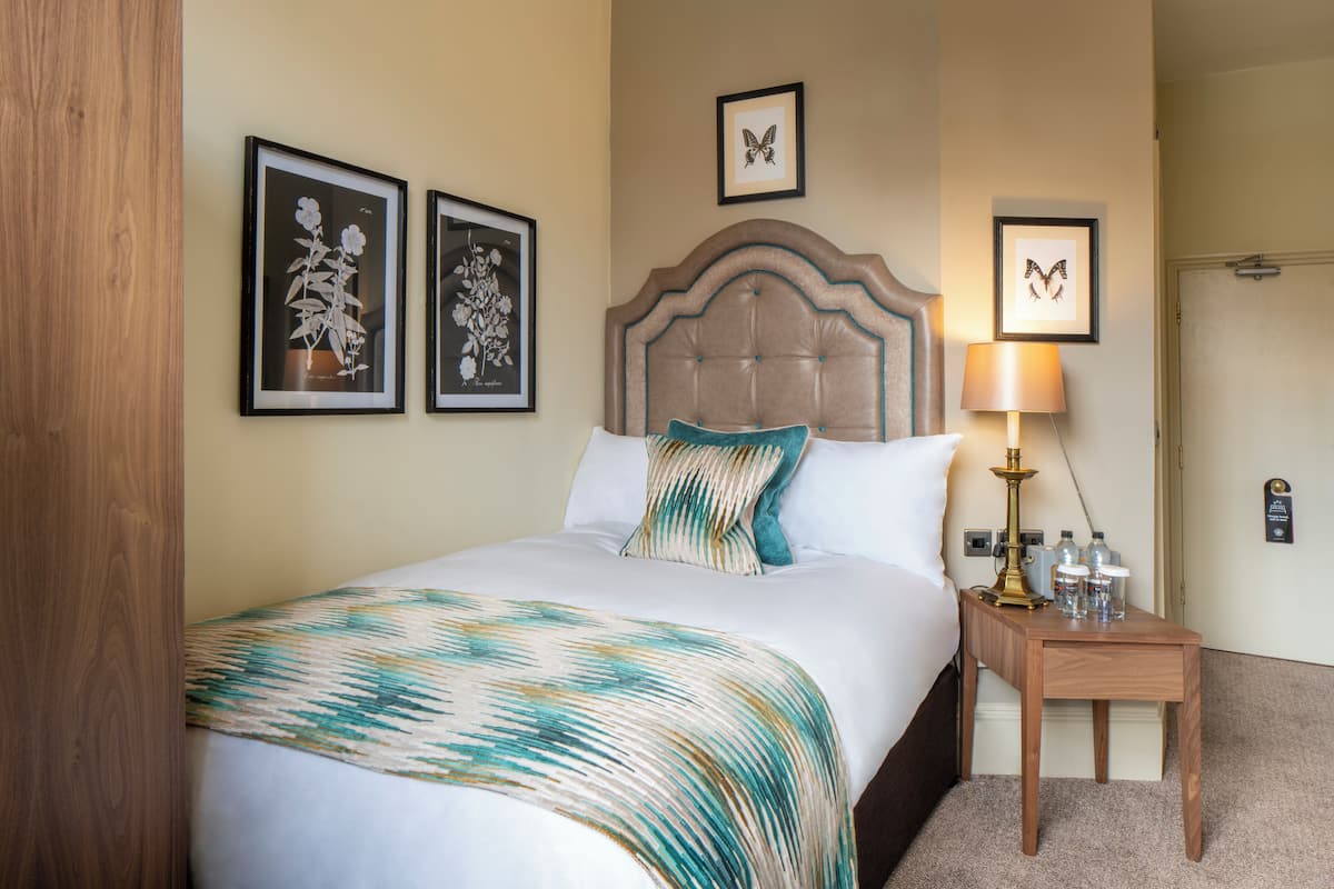 Een net opgemaakt bed met een bedsprei met patroon, een houten nachtkastje met een lamp, en ingelijste botanische prints aan de muur.