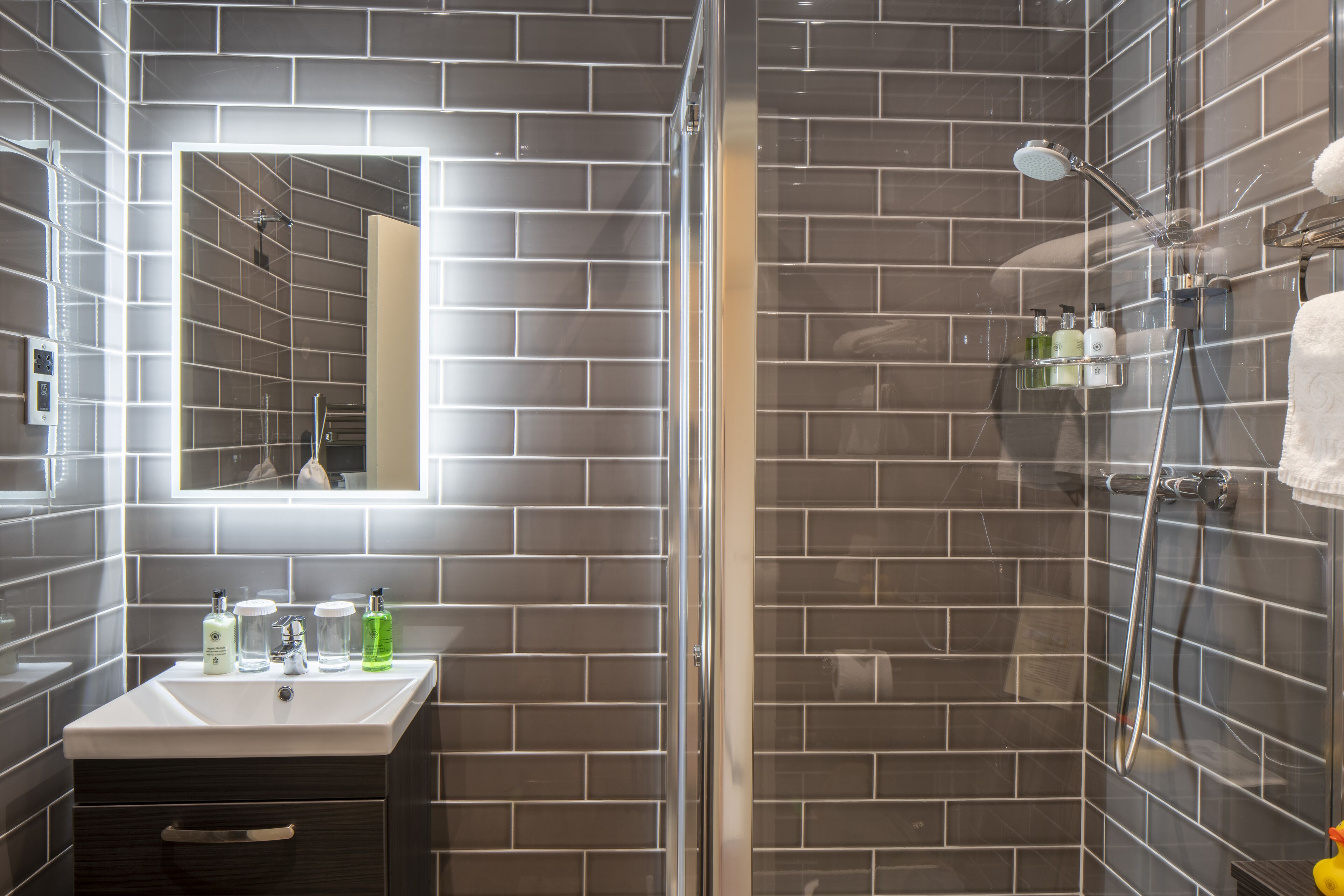 A modern bathroom with a glass-enclosed shower, a vanity with a mirror, and toiletries on the sink.