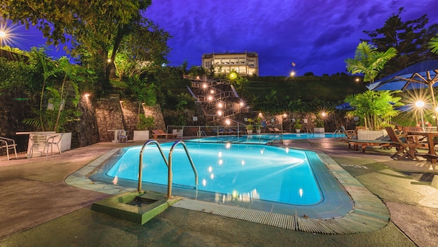 A swimming pool with a view of a building on a hill, surrounded by trees and outdoor seating.