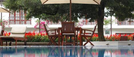 A poolside area with a table and chairs under a large umbrella, surrounded by lush greenery and a clear pool reflecting the environment.