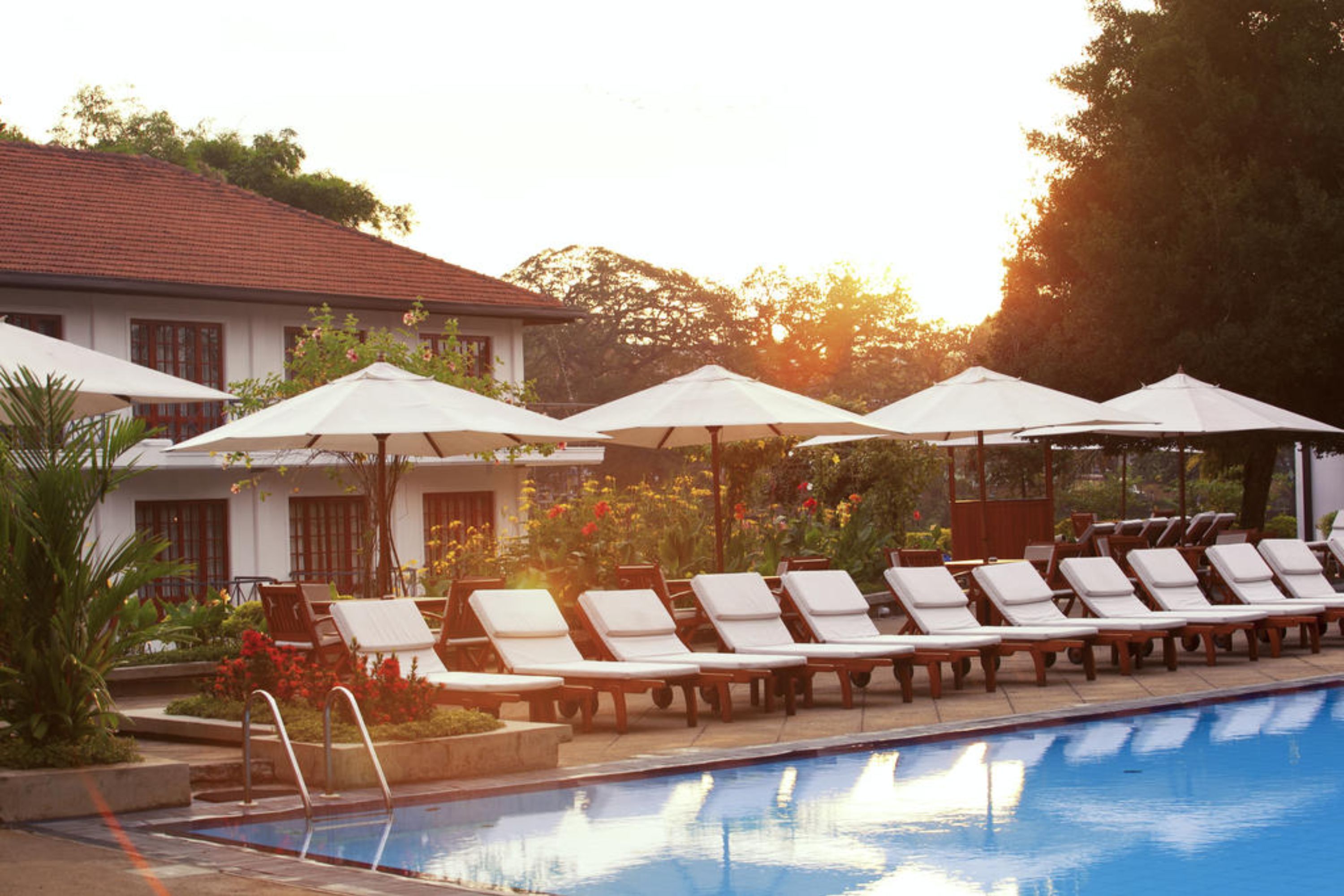 Un espace piscine doté de chaises longues et de parasols, entouré d’un bâtiment avec un toit en tuiles et d’un jardin fleuri.