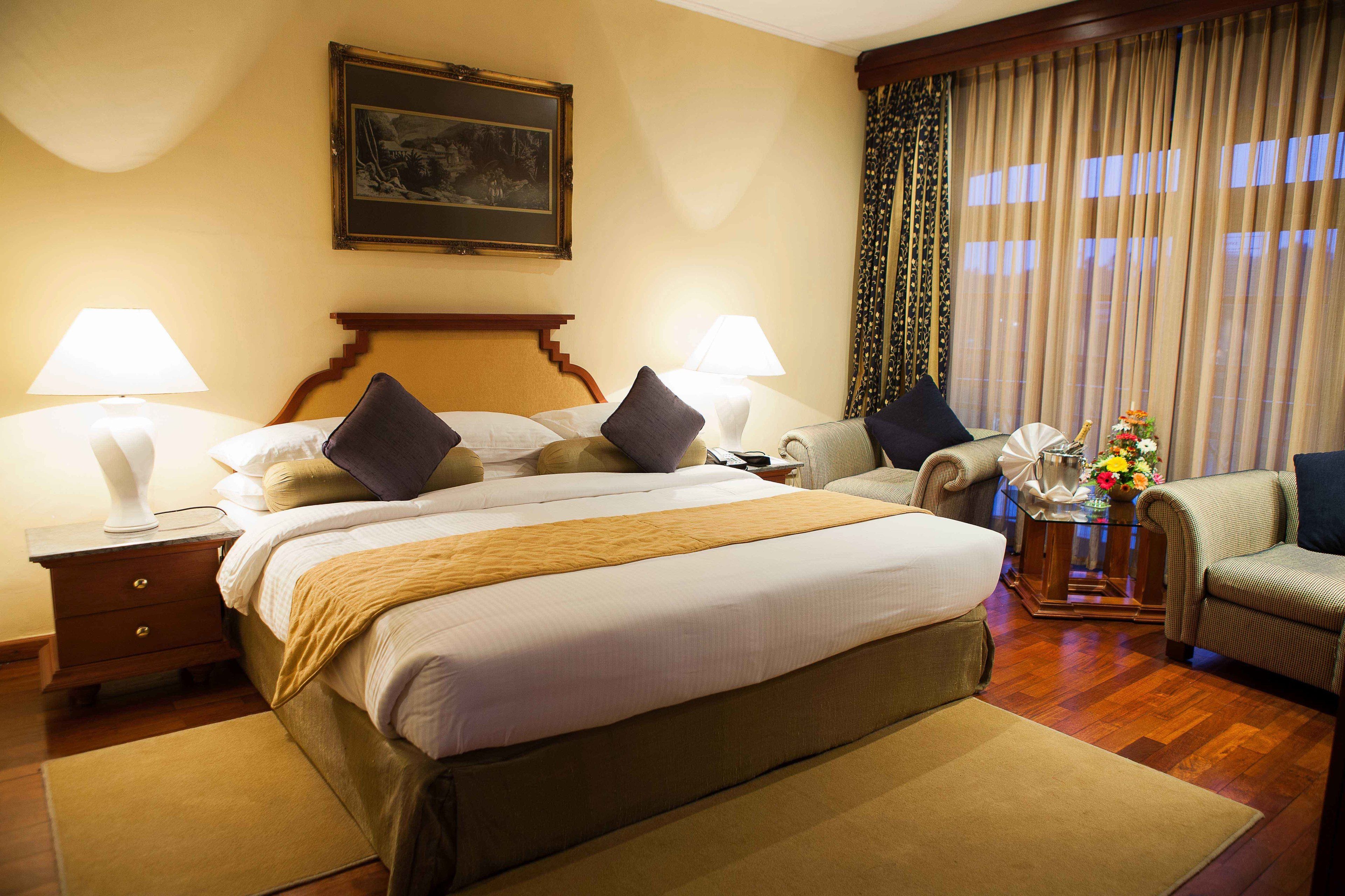 A hotel room with a large bed, two bedside tables with lamps, a chair, a small table with a vase of flowers, and a painting on the wall.