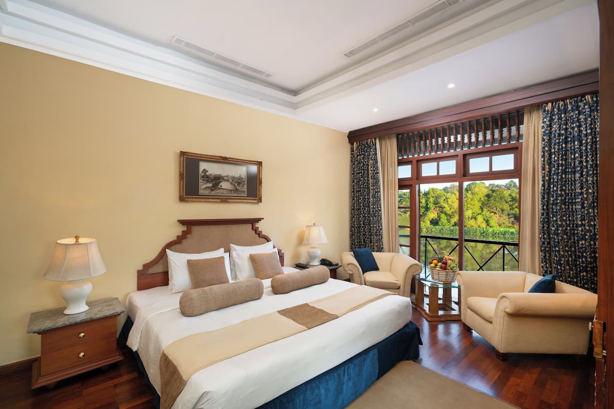 Habitación de hotel con una cama grande, dos mesitas de noche, una silla, una pequeña mesa con una cesta de frutas y vistas a la vegetación por la ventana.