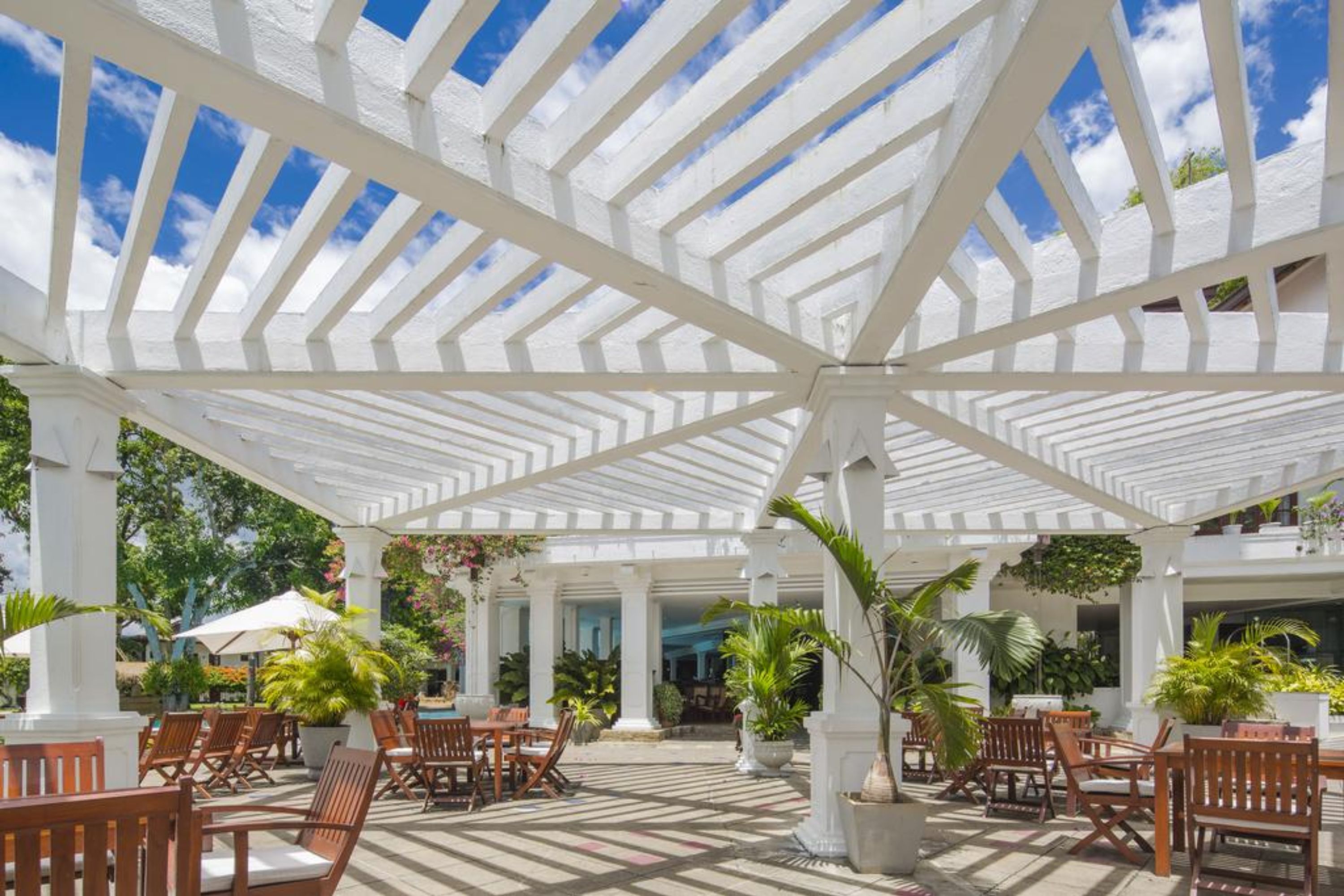 A covered outdoor seating area with wooden chairs and tables, surrounded by potted plants and a white pergola structure.