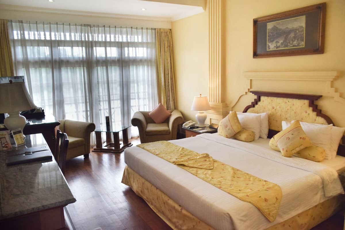 Habitación de hotel con una cama grande, un escritorio con lámpara, una silla y un ventanal con cortinas.