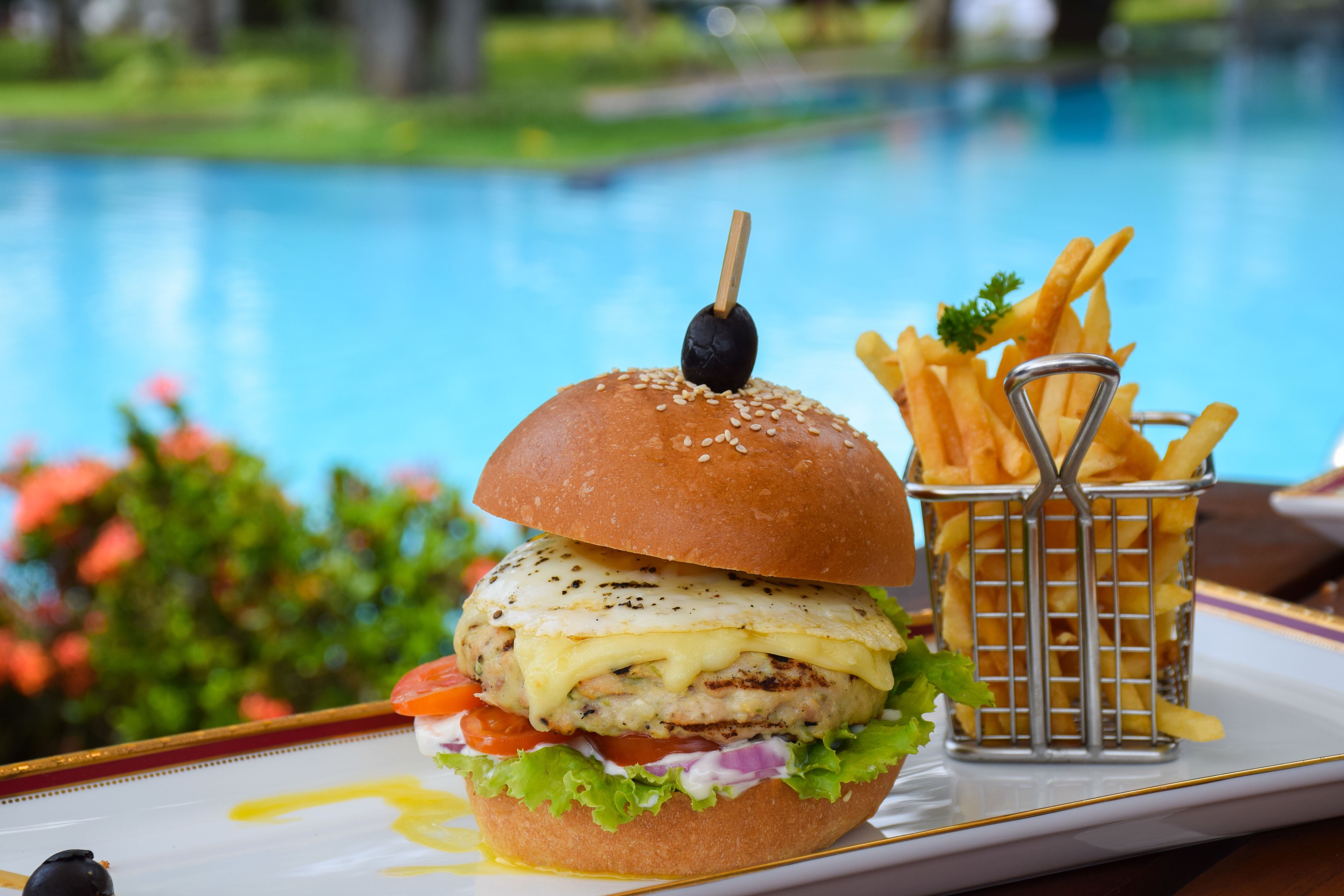 A burger with a sesame seed bun, grilled chicken, cheese, lettuce, tomato, and a black olive on a toothpick, accompanied by a basket of French fries.