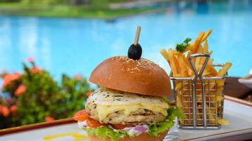 A burger with a sesame seed bun, grilled chicken, cheese, lettuce, tomato, and a black olive on a toothpick, accompanied by a basket of French fries.