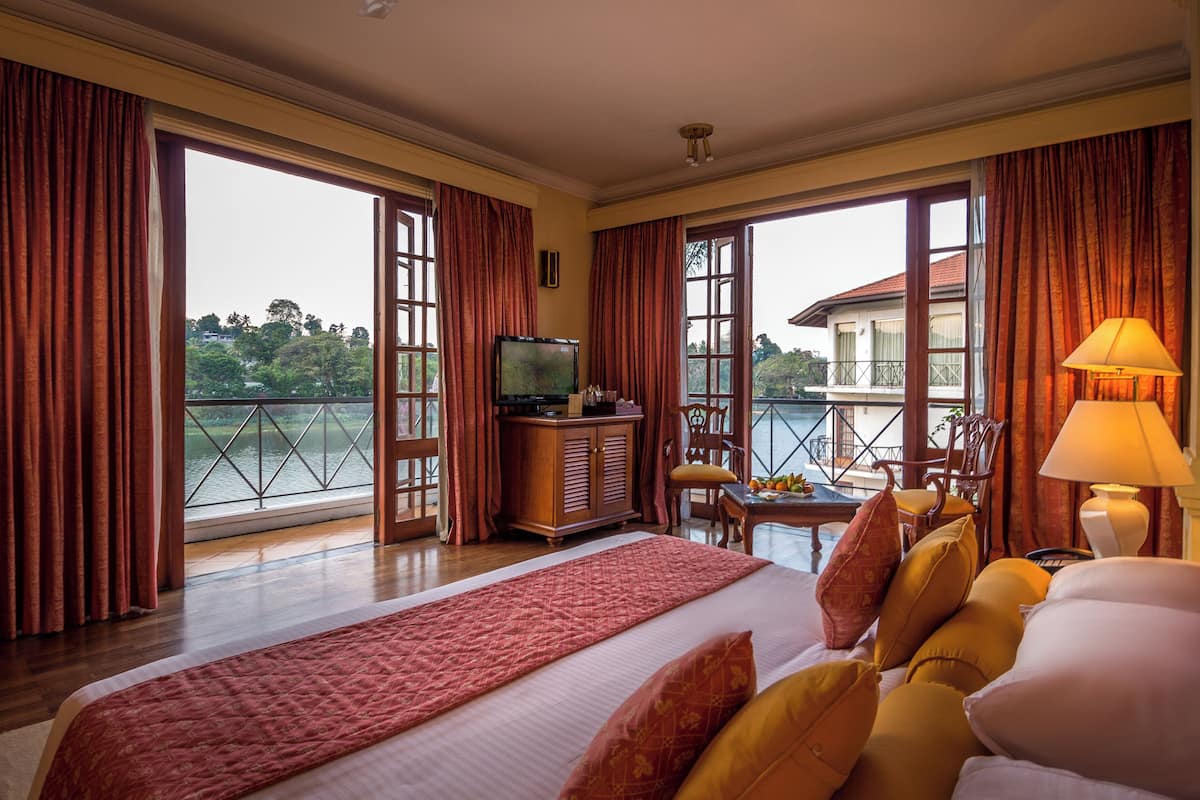 Habitación de hotel con una cama grande, televisión, escritorio con lámpara y vista a un lago a través de amplias ventanas.