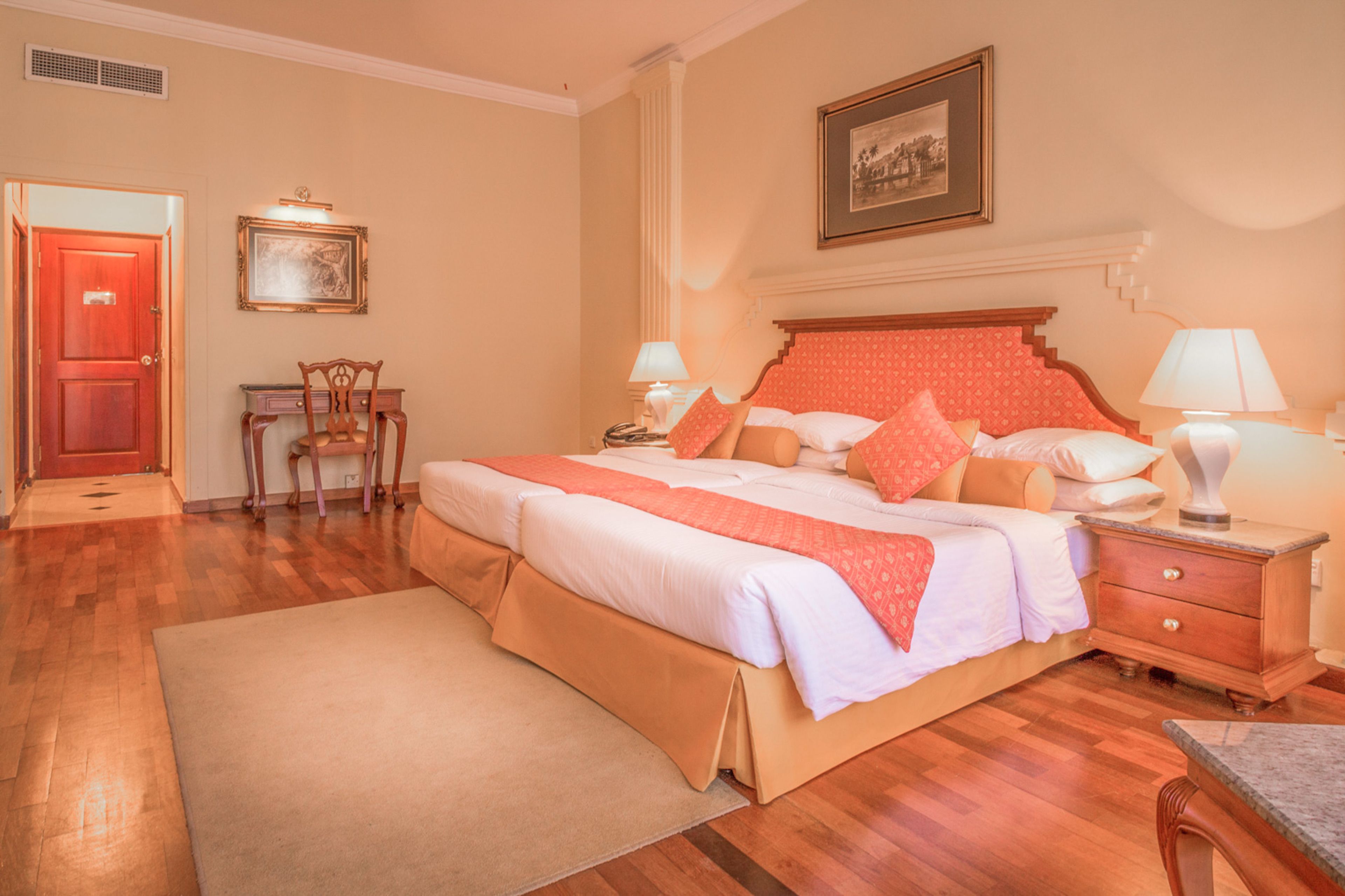 Quarto de hotel com uma cama grande, duas mesas de cabeceira com luminárias, uma cadeira de madeira, uma escrivaninha e uma porta.