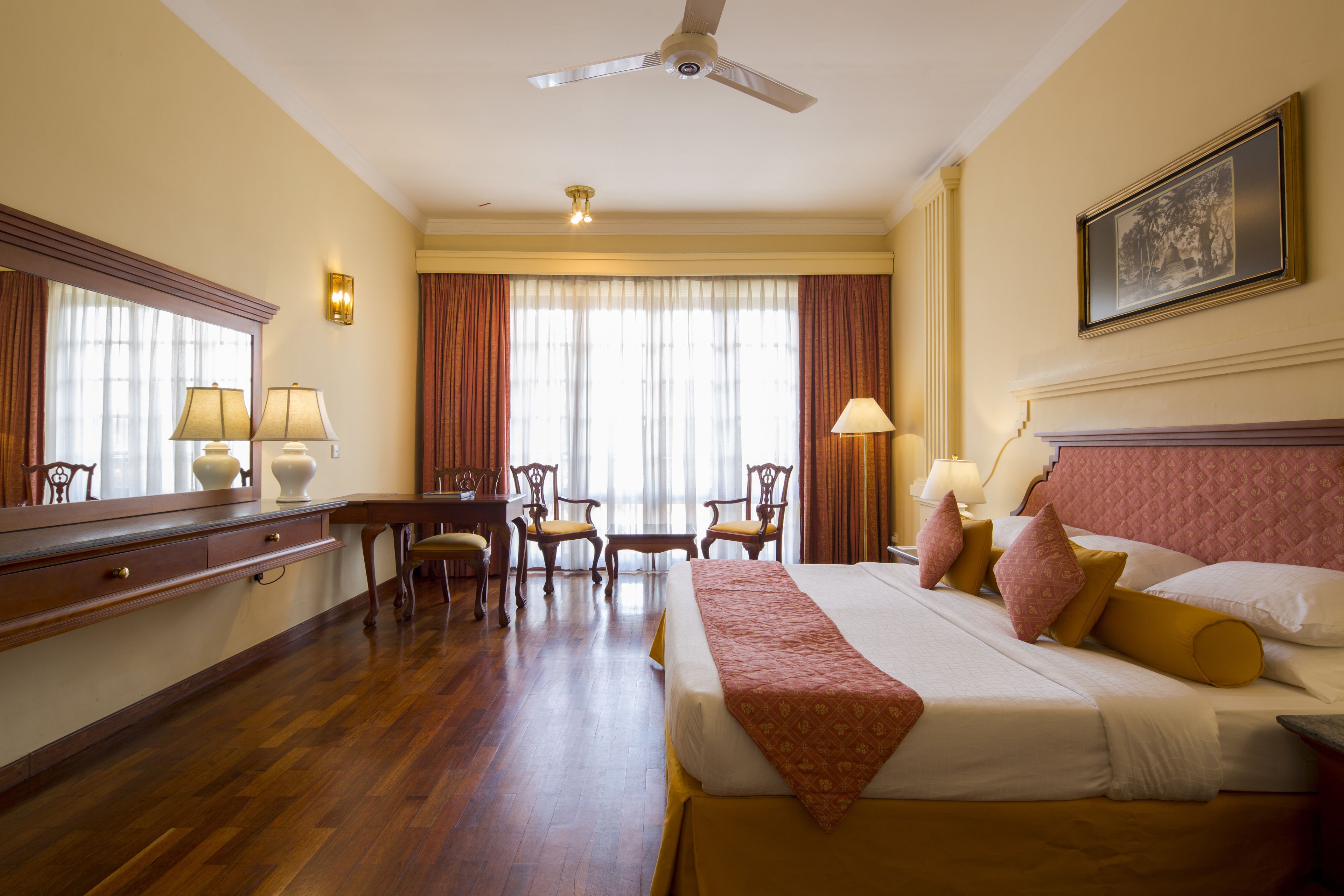A hotel room with a large bed, a desk with a chair, a vanity with a mirror, and a ceiling fan.