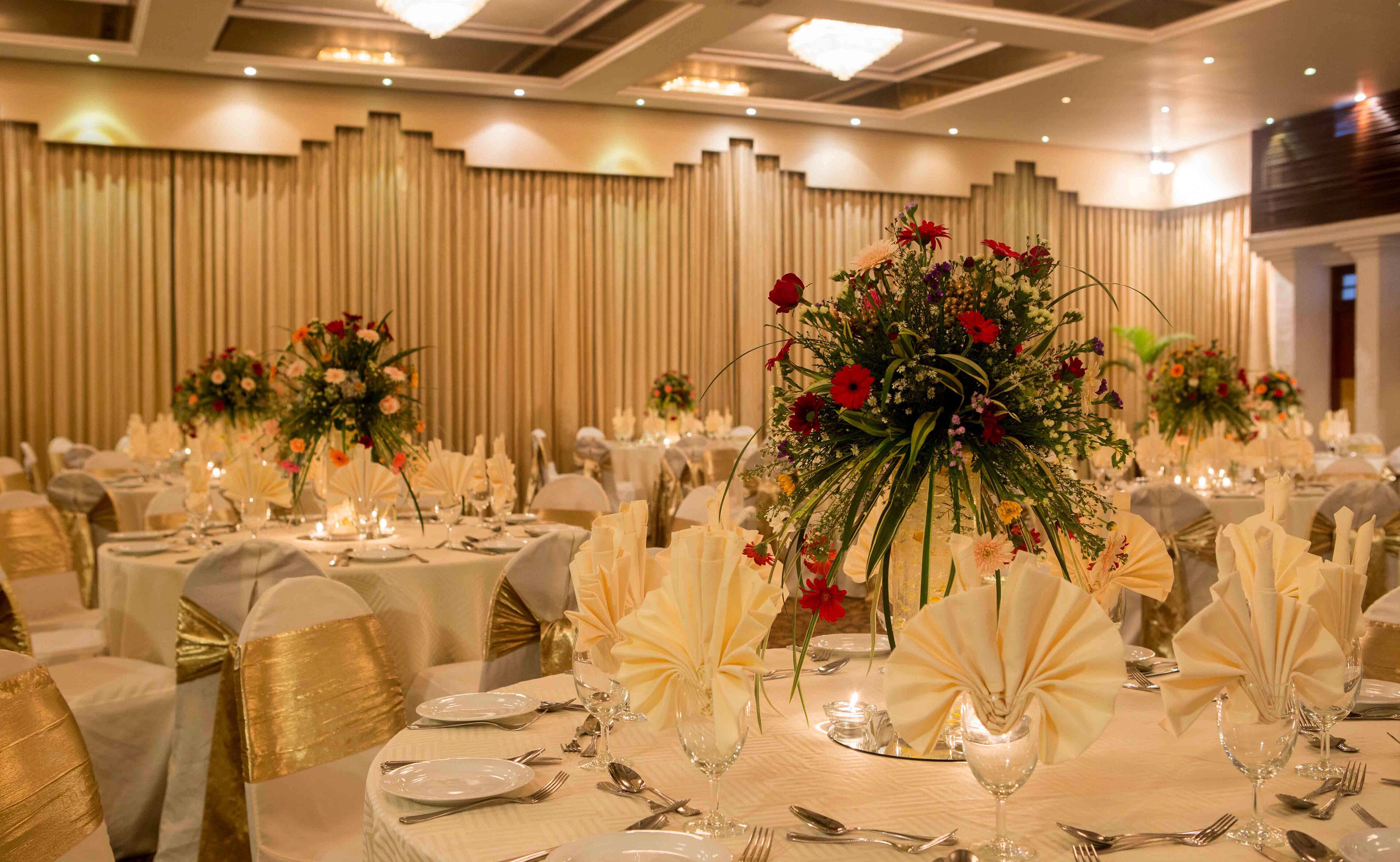 Un salón de banquetes con mesas redondas dispuestas para un evento formal, con manteles blancos, fajones dorados y centros de mesa florales.