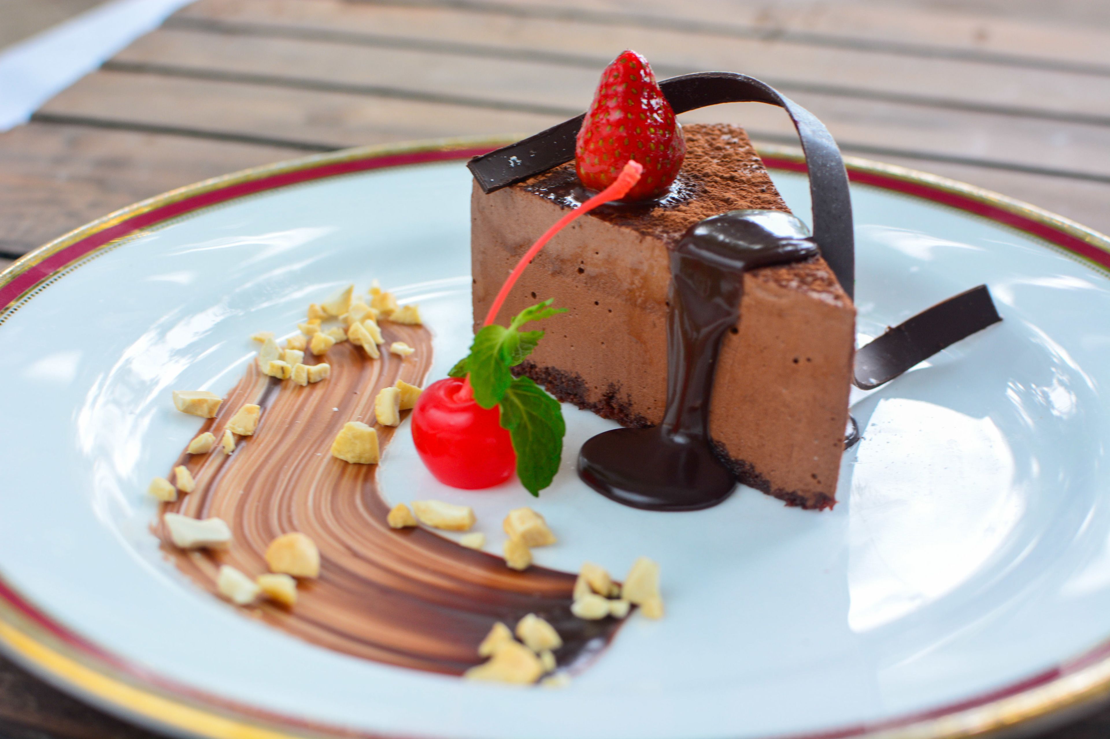Una porción de pastel de chocolate con una fresa encima, decorada con salsa de chocolate y nueces, sobre un plato blanco con borde rojo.