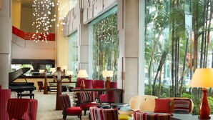 A spacious lobby with a grand piano, red upholstered chairs, and a large window offering a view of palm trees. - Grand Mercure Medan Angkasa (Medan)